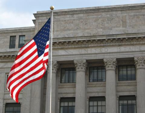 An American flag on a pole in front of a stone building with columns, featuring the words "UNITED STATES" carved above the entrance.